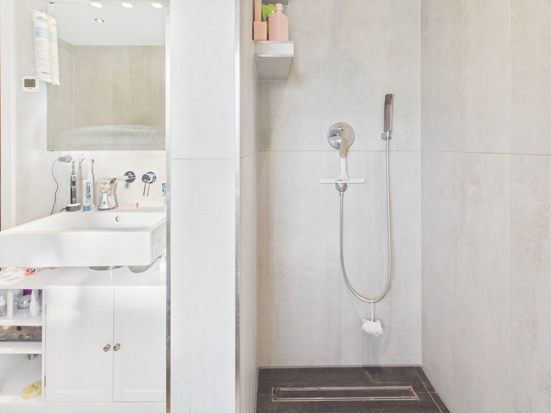 Modernes Badezimmer mit Dusche, Waschbecken und Spiegel. Helle Fliesen und Regale mit Toilettenartikeln.