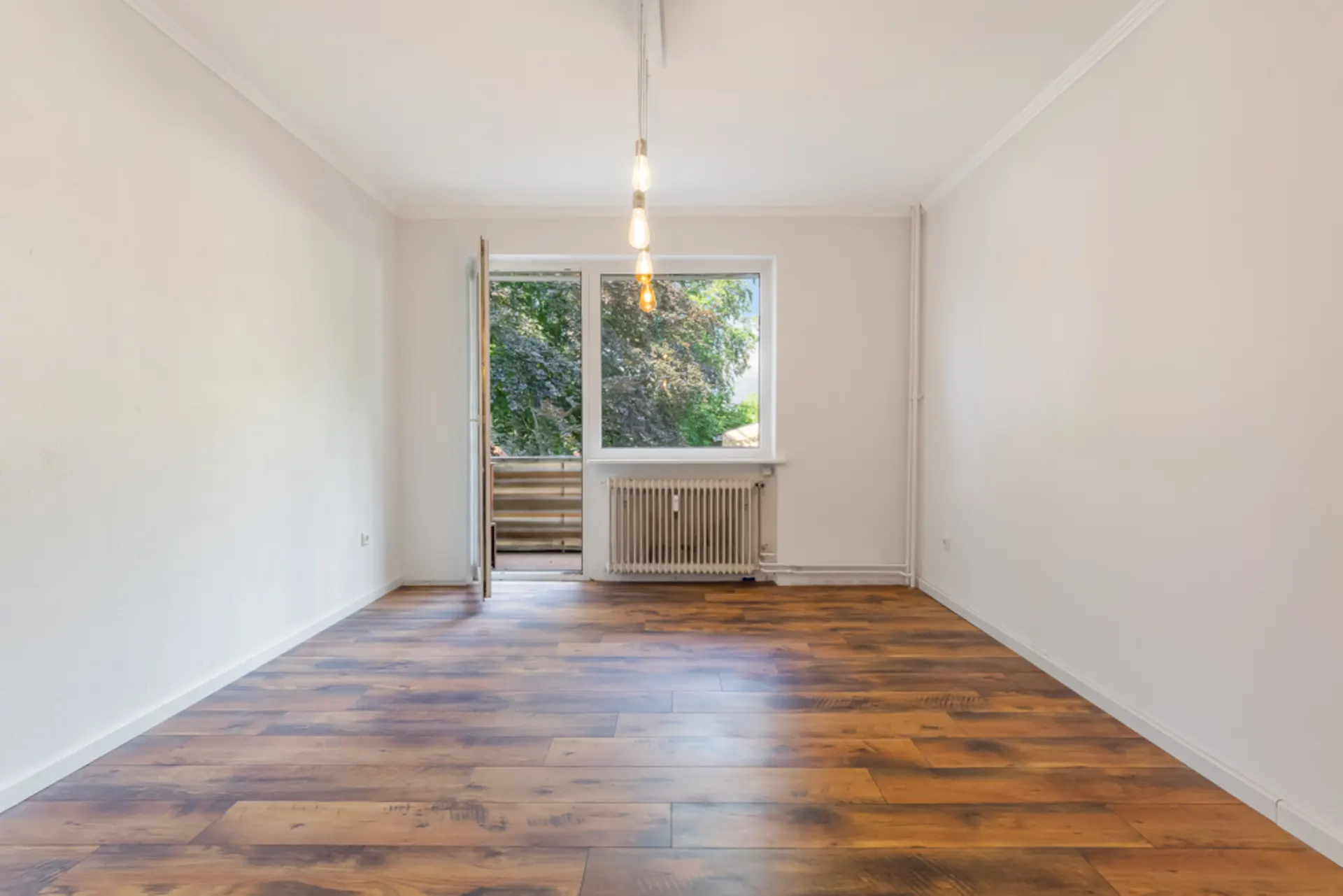 Leerer Raum mit Holzfußboden, großem Fenster und Balkontür, Blick ins Grüne.