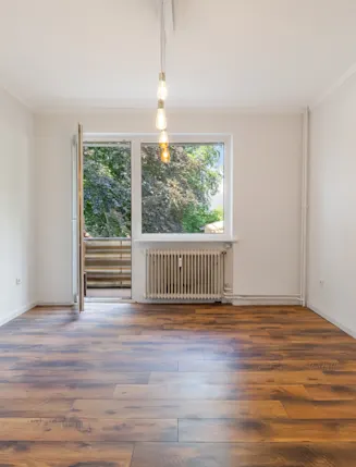 Leerer Raum mit Holzfußboden, großem Fenster und Balkontür, Blick ins Grüne.