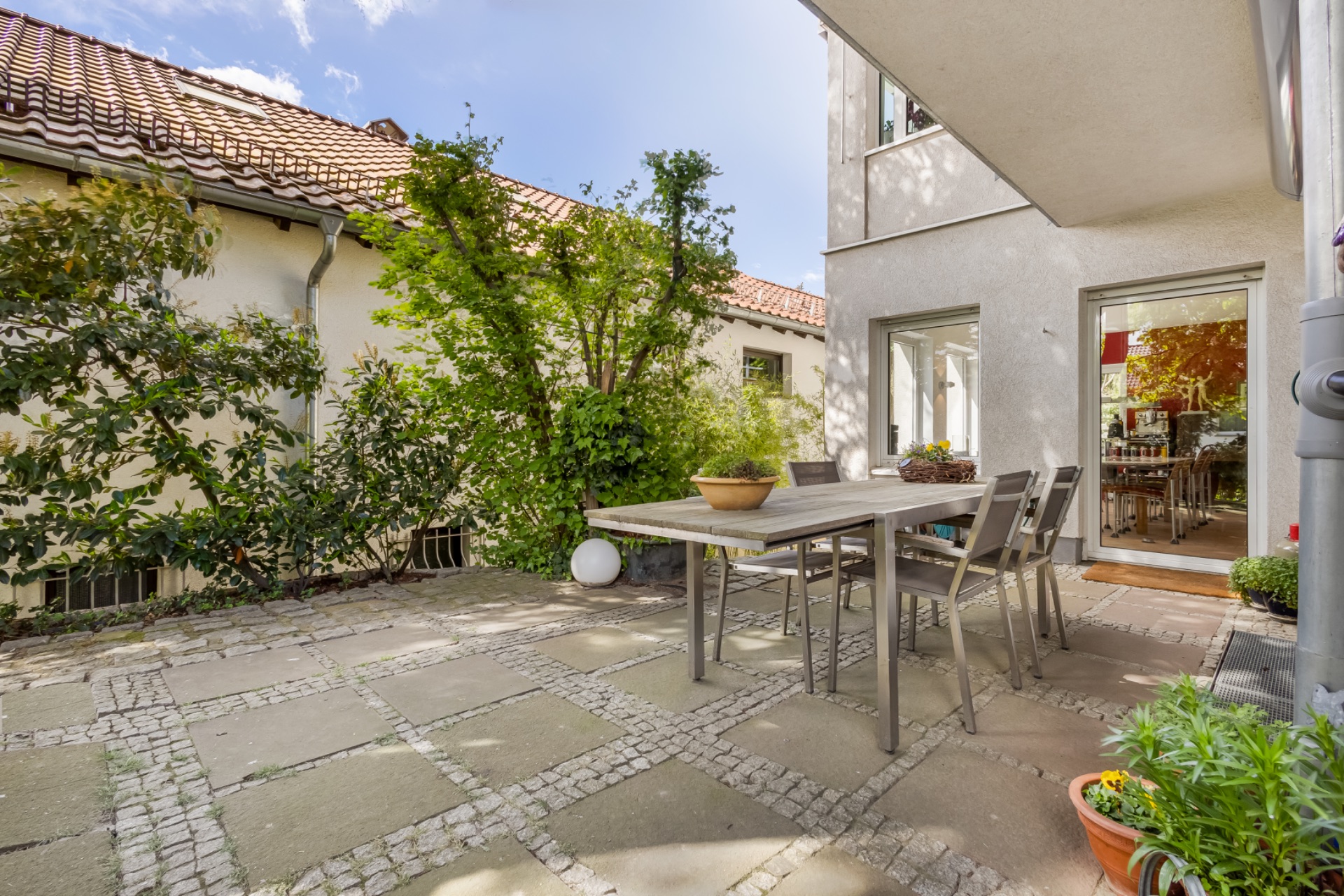 Gepflasterte Terrasse mit Esstisch und Stühlen, umgeben von Pflanzen und Büschen neben einem Wohnhaus.