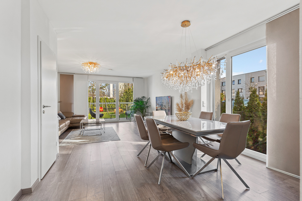 Helles Esszimmer mit großem Tisch, sechs Stühlen und Kronleuchter, im Hintergrund Wohnbereich mit Sofa.