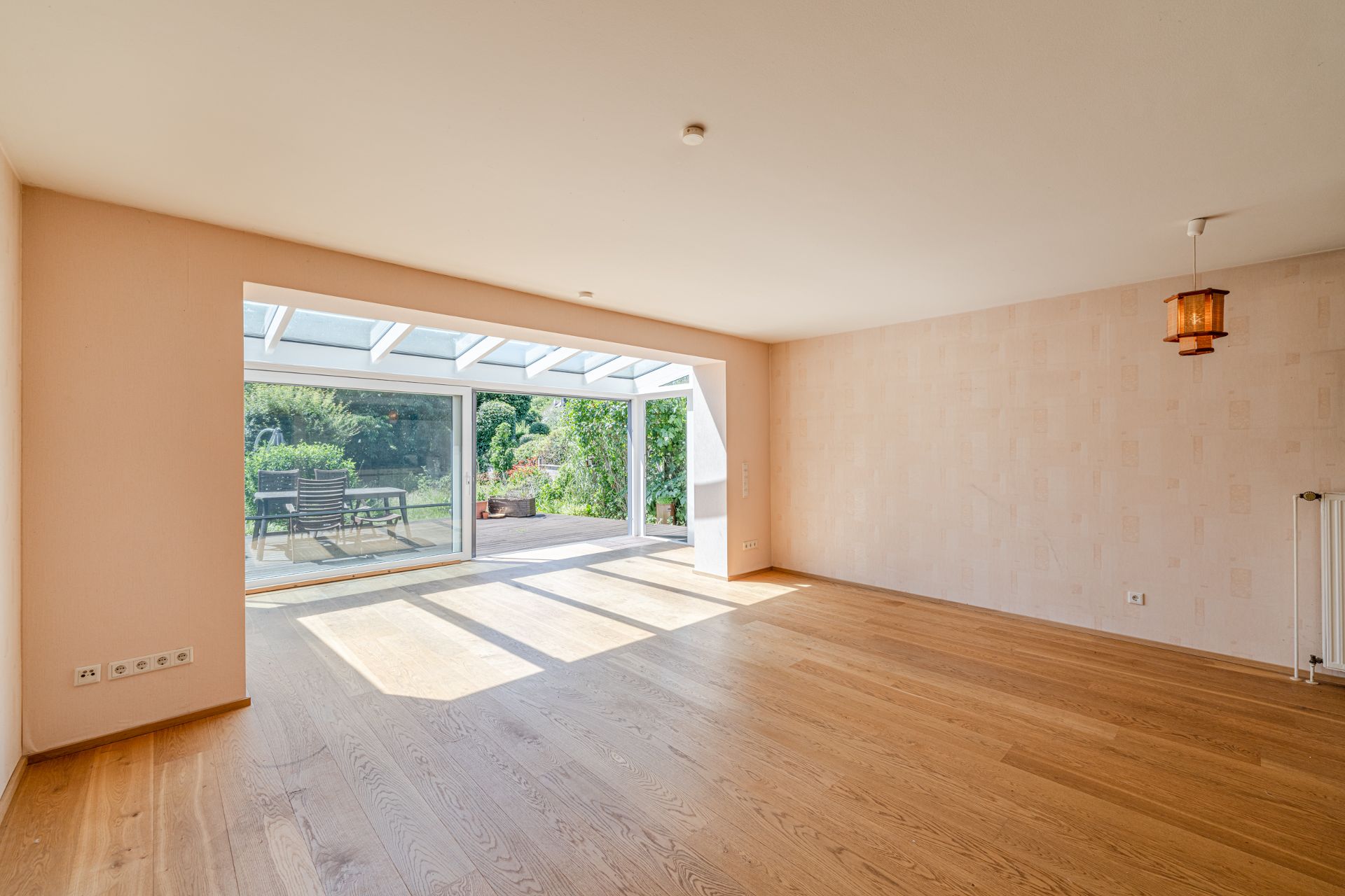Helles Wohnzimmer mit Holzfußboden, großem Fenster und Zugang zur Terrasse mit Gartenmöbeln.
