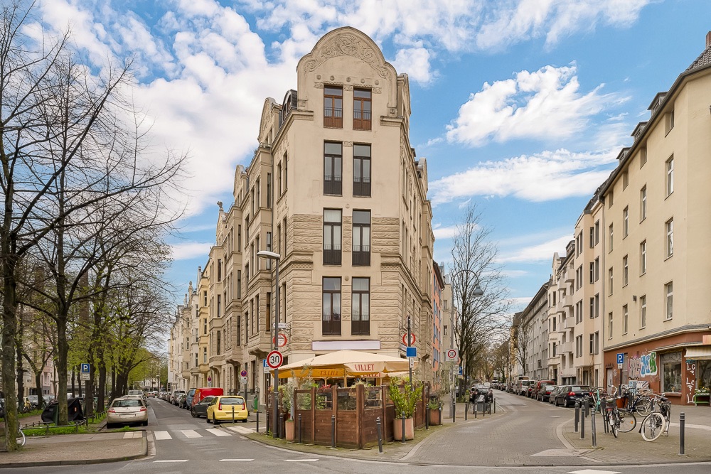 Eckhaus mit mehreren Etagen, Cafégarten im Erdgeschoss, umgeben von Straßen und geparkten Autos.
