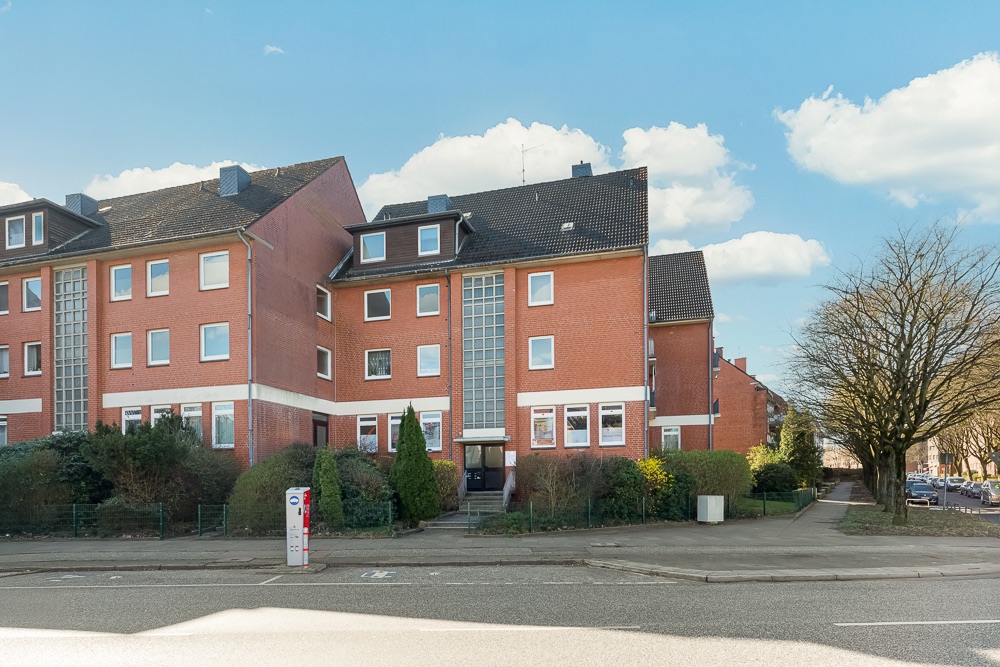 Mehrfamilienhaus aus rotem Backstein mit mehreren Etagen und Fenstern, davor Büsche und Gehweg.