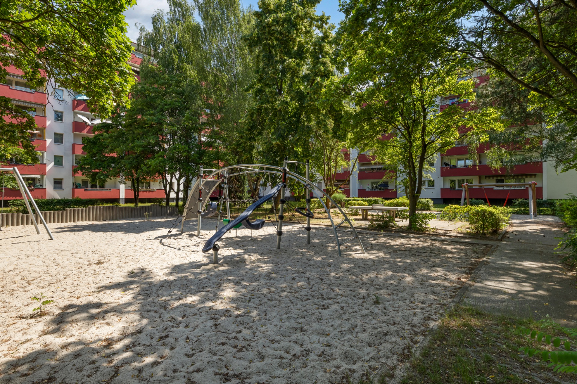 Spielplatz mit Klettergerüst und Sandfläche, umgeben von Bäumen und Wohngebäuden.
