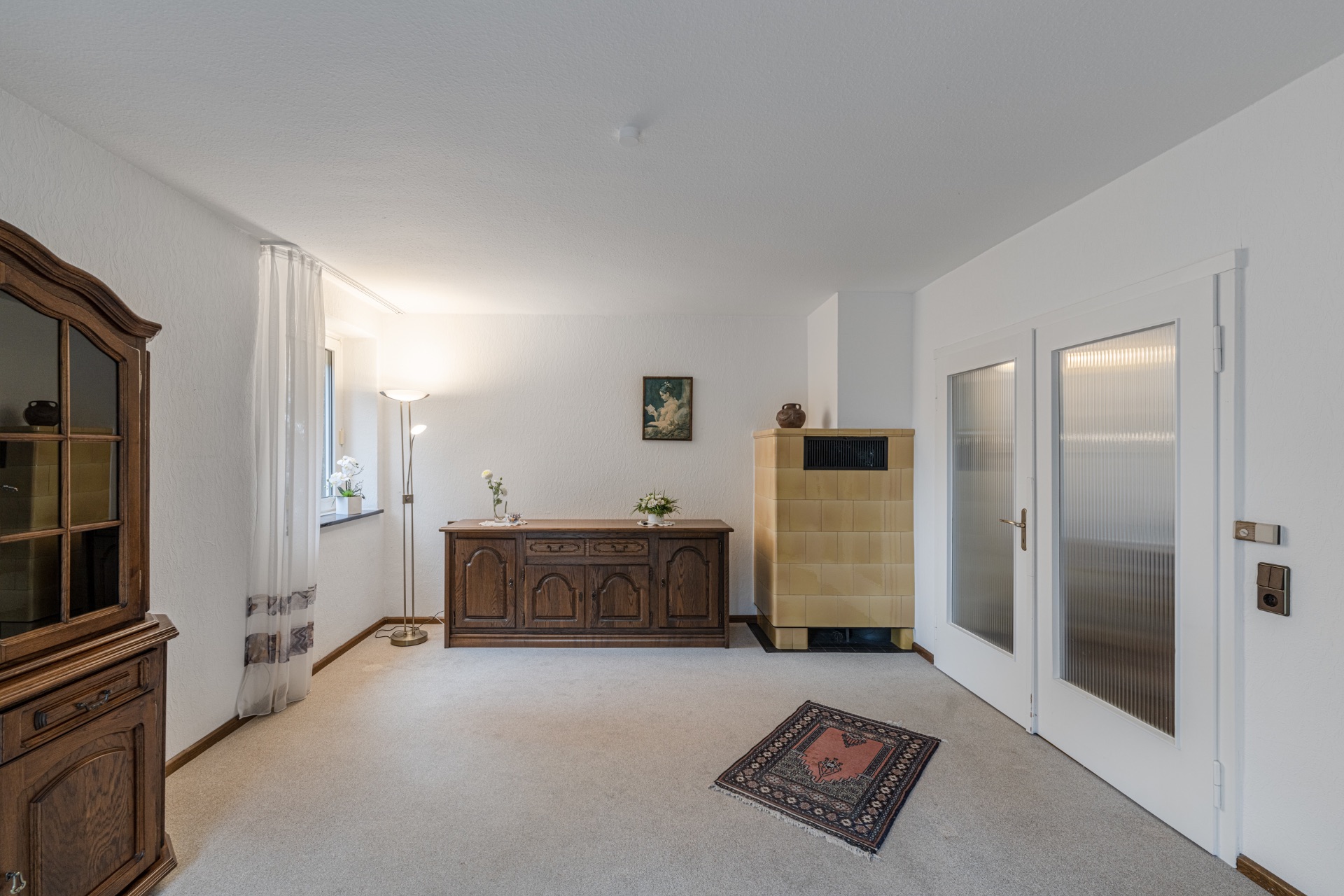 Wohnzimmer mit Teppich, Holzmöbeln, Stehlampe und gelbem Kachelofen.