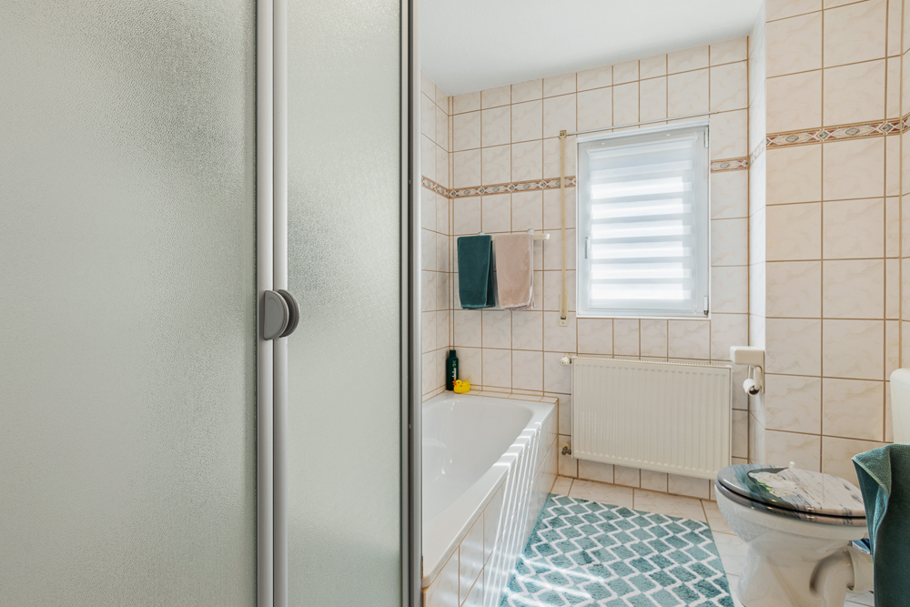 Gefliestes Badezimmer mit Dusche, Badewanne, WC, Fenster und Handtüchern an der Wand.