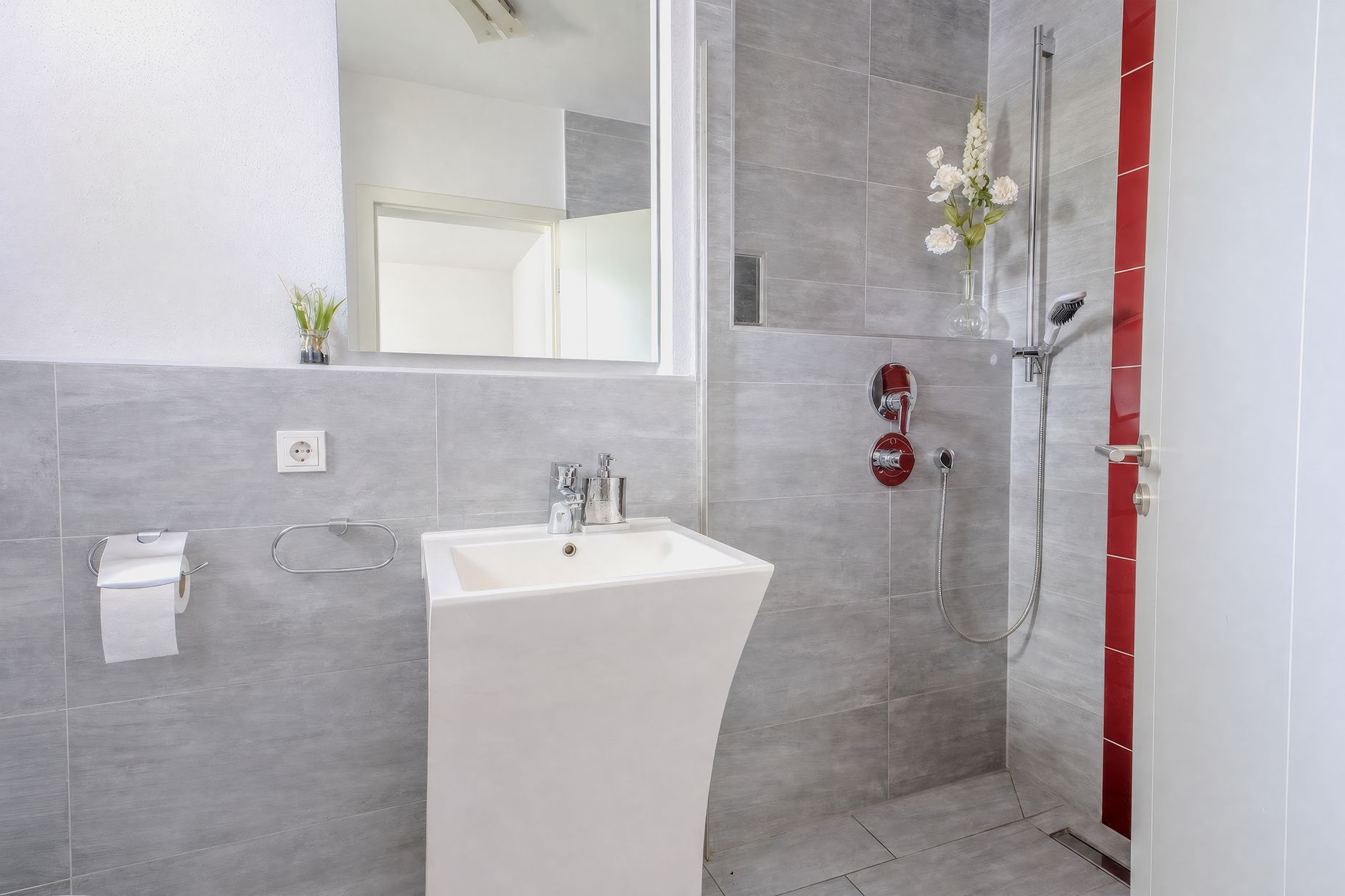 Modernes Badezimmer mit Waschbecken, Spiegel und ebenerdiger Dusche, graue Fliesen und rote Akzente.