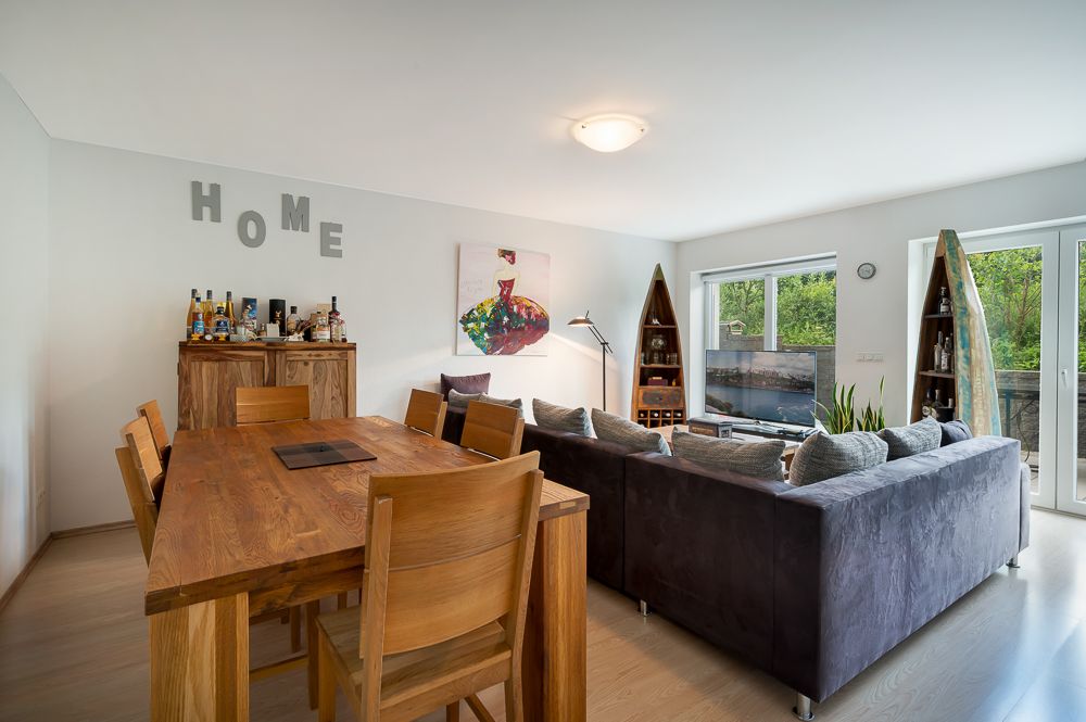 Wohnzimmer mit großem Holztisch, grauem Sofa und Regalen, viel Tageslicht durch Fenster.