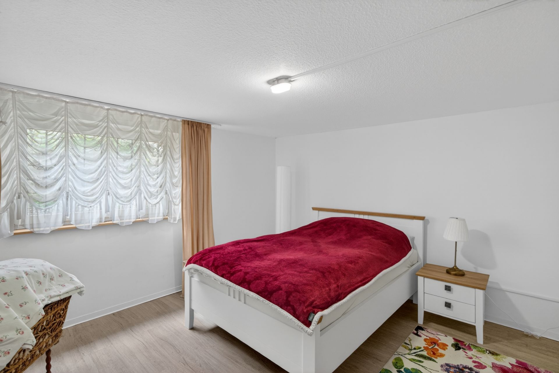Schlafzimmer mit weißem Bett, roter Decke, Nachttisch und Lampe, hellem Vorhang am Fenster.