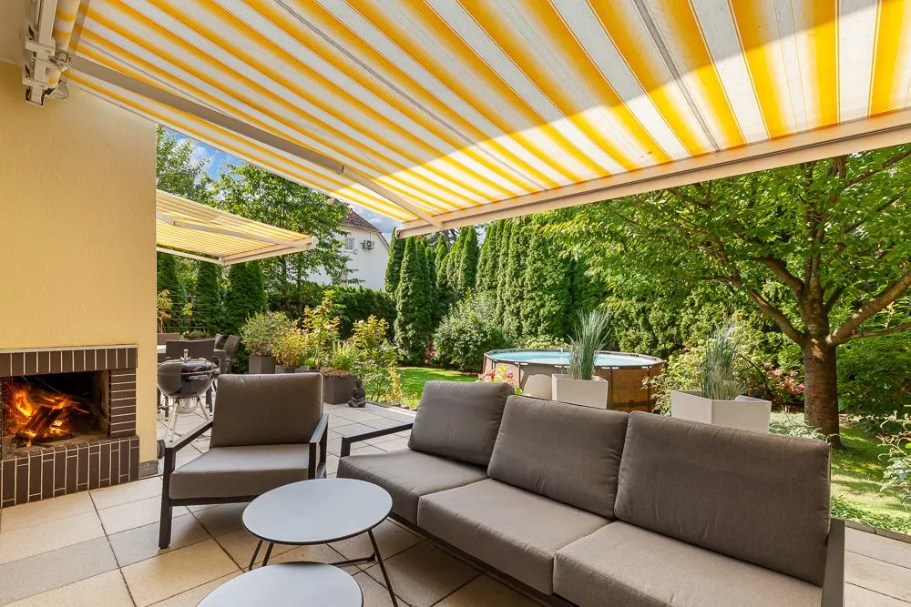 Terrasse mit Markise, Sofas, runden Tischen, Kamin und Blick auf Garten mit Whirlpool.