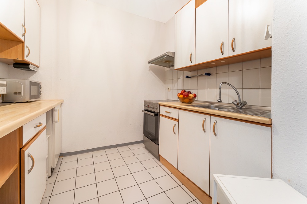 Kleine Küche mit weißen Schränken, Spüle, Herd, Mikrowelle und Obstschale auf der Arbeitsplatte.