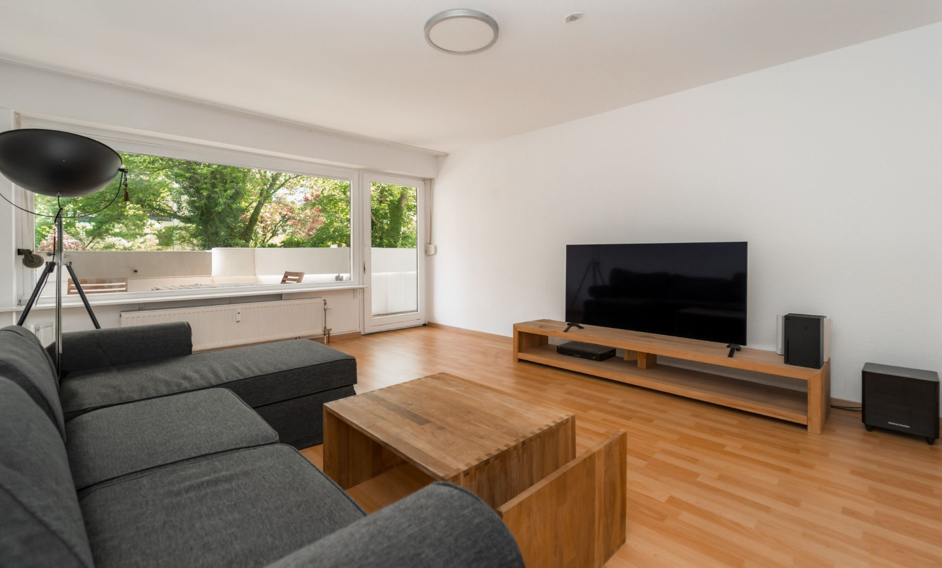 Wohnzimmer mit grauem Sofa, Holztisch, Fernseher und großem Fenster mit Blick ins Grüne.