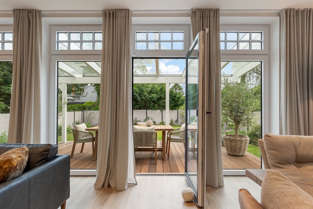 Große Glas-Schiebetür mit Vorhängen, Blick auf Terrasse mit Sitzmöbeln und Garten.