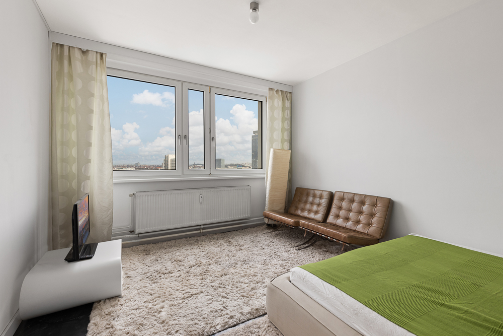 Helles Schlafzimmer mit großem Fenster, braunem Sofa, grünem Bett und Teppich.