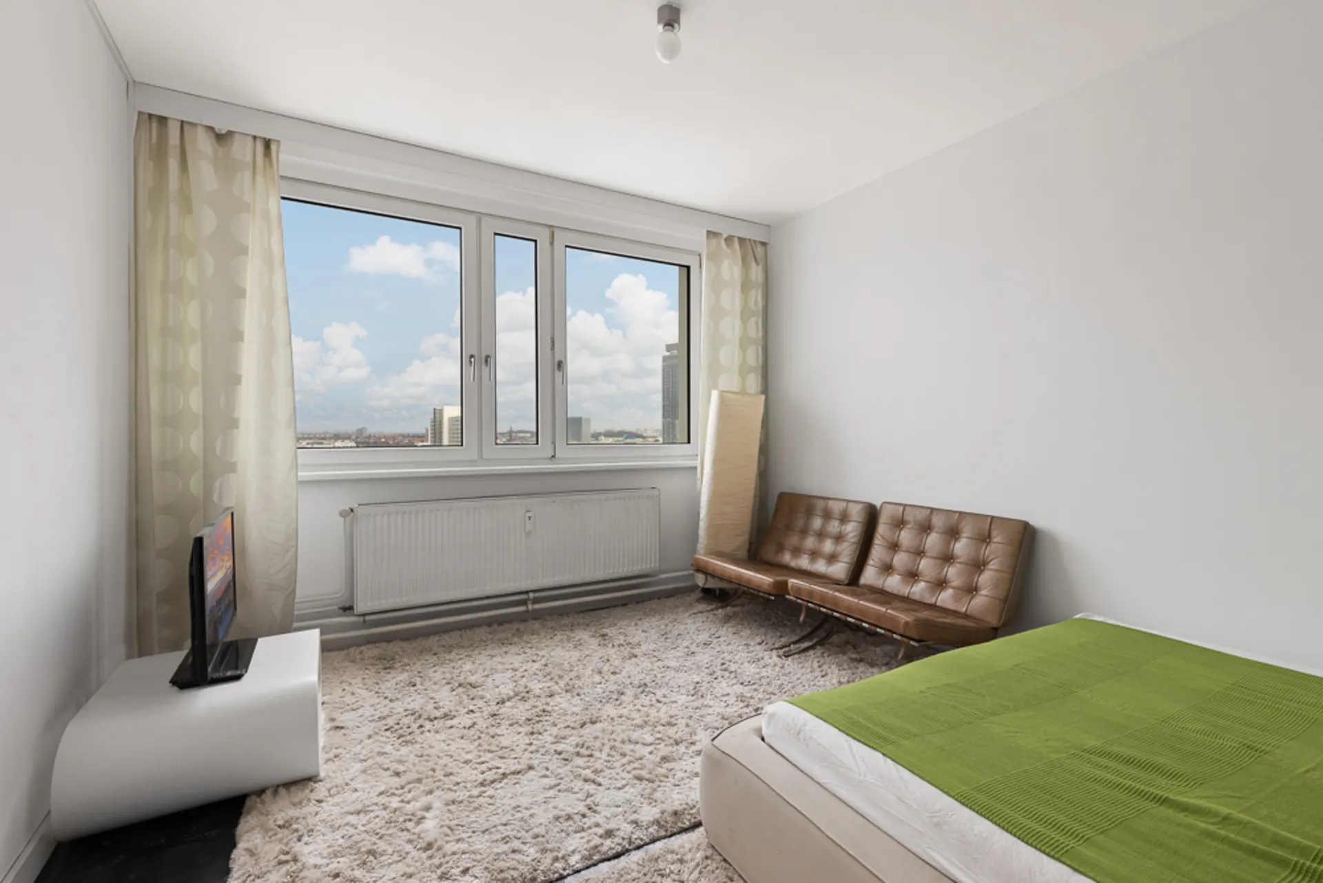 Helles Schlafzimmer mit großem Fenster, braunem Sofa, grünem Bett und Teppich.