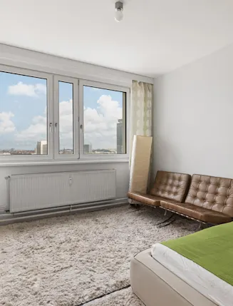 Helles Schlafzimmer mit großem Fenster, braunem Sofa, grünem Bett und Teppich.