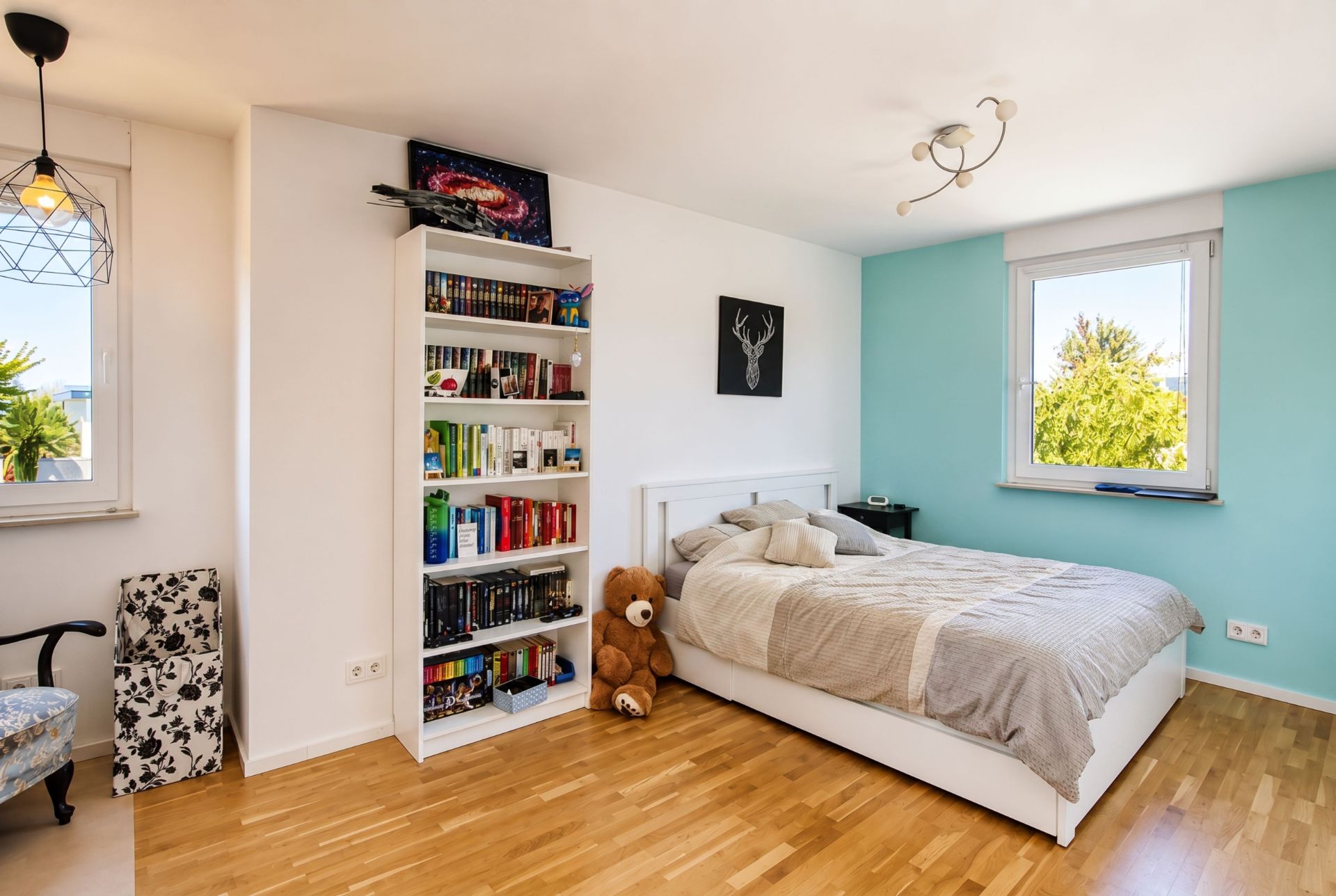 Helles Schlafzimmer mit Bett, Bücherregal, großem Teddybär und Fenster mit Ausblick.