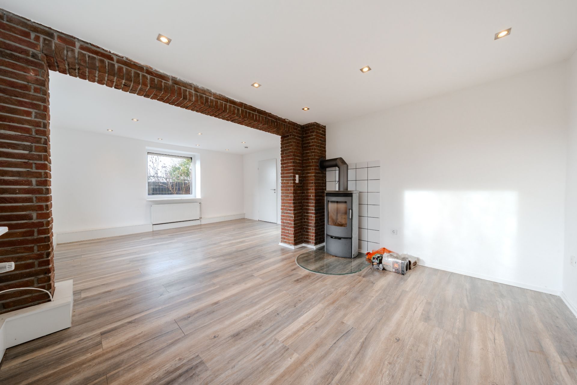 Helles Wohnzimmer mit Holzofen, Ziegelbogen und großem Fenster, moderner Holzboden.