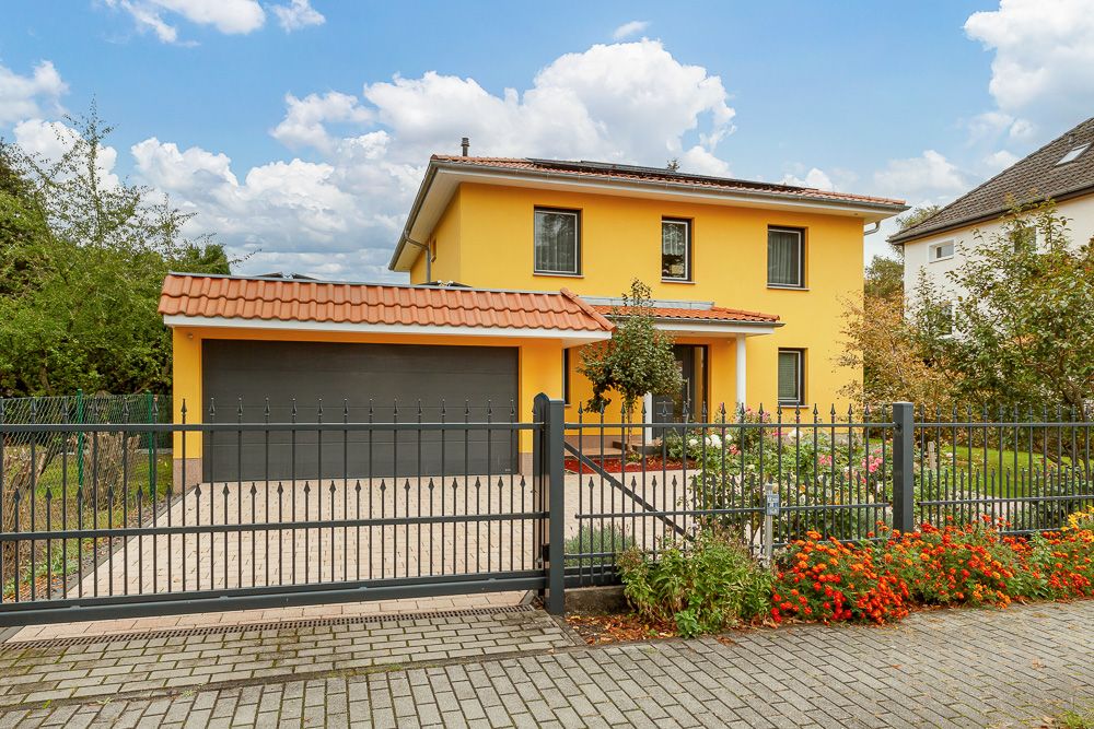 Zweistöckiges gelbes Wohnhaus mit Garagentor, Garten und schwarzem Zaun vor dem Haus.