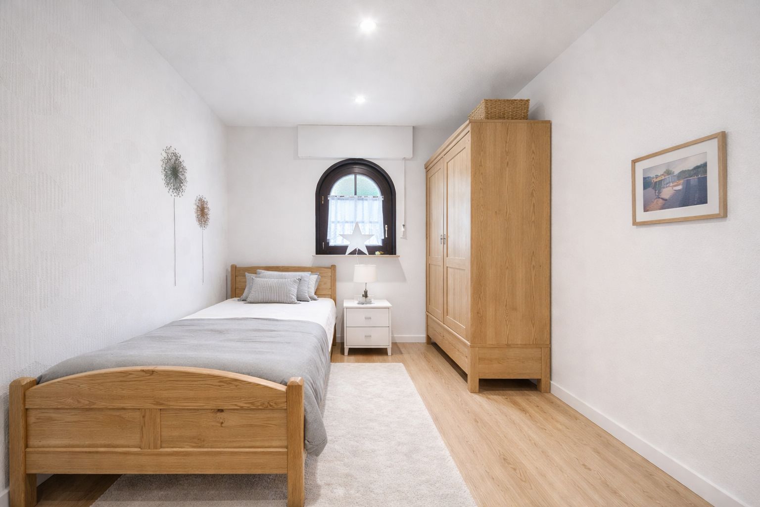 Helles Schlafzimmer mit Einzelbett, Holzschrank, Nachttisch und Fenster mit Rundbogen.