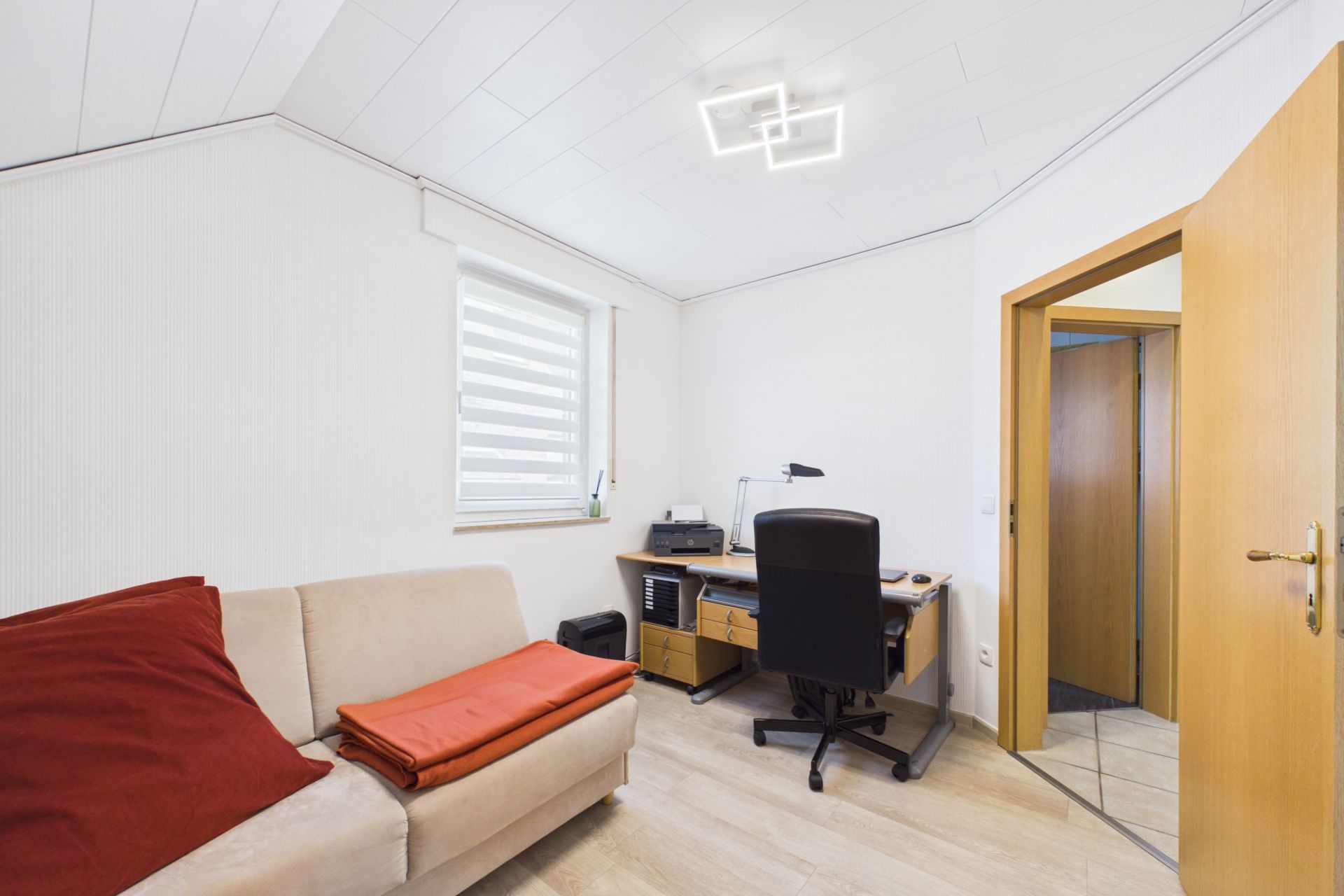 Helles Arbeitszimmer mit Schreibtisch, Bürostuhl, Sofa und offenem Türrahmen.