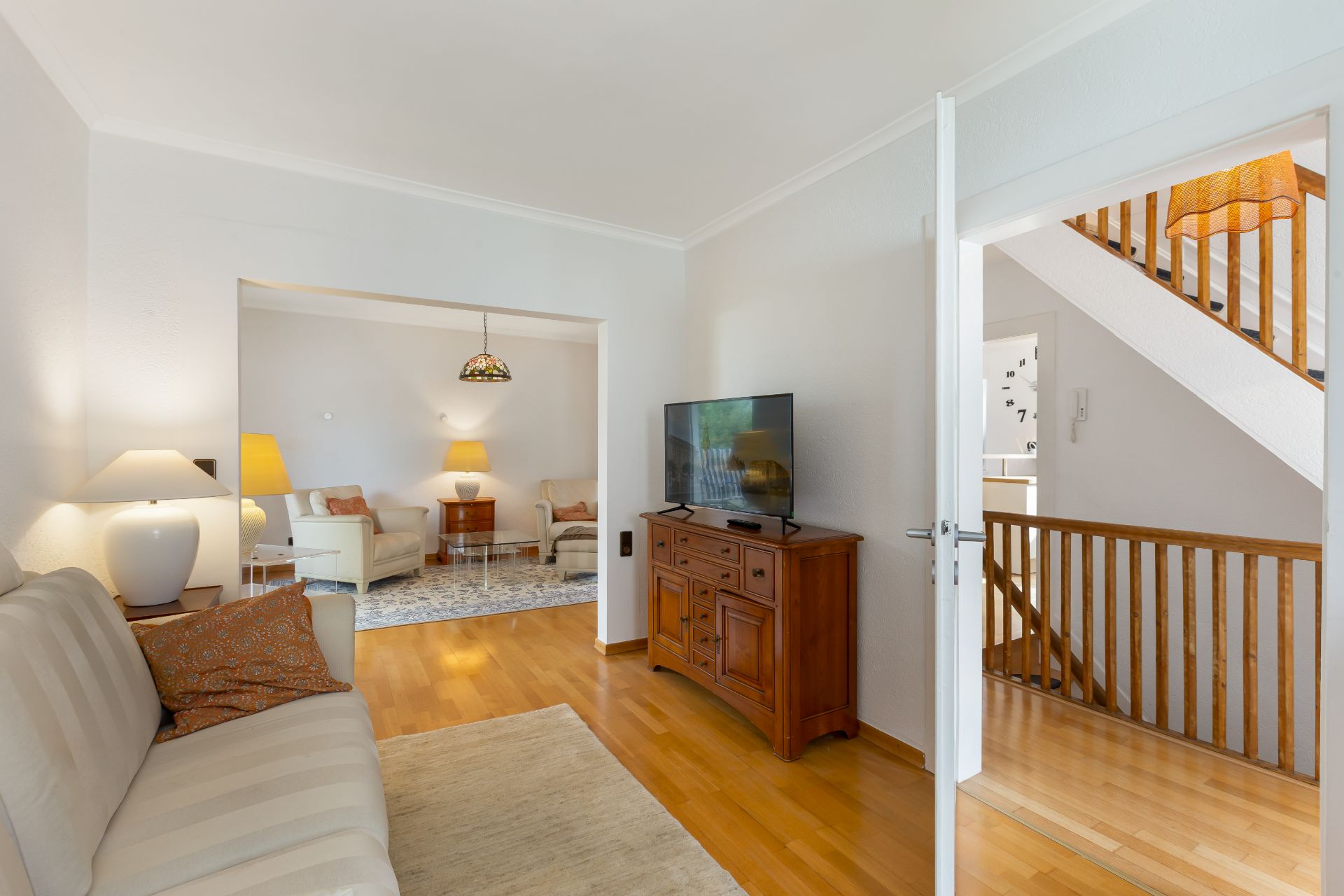 Helles Wohnzimmer mit Sofa, Sessel, TV auf Holzkommode und Treppe im Hintergrund.