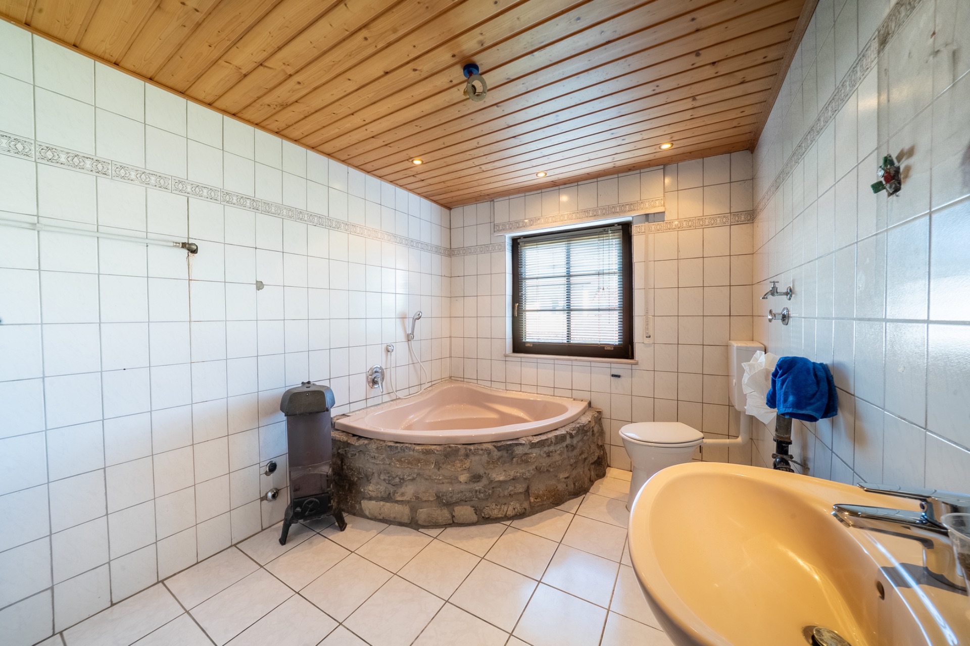 Badezimmer mit Eckbadewanne aus Stein, Waschbecken, WC und Fenster, Holzdecke und weiße Fliesen.