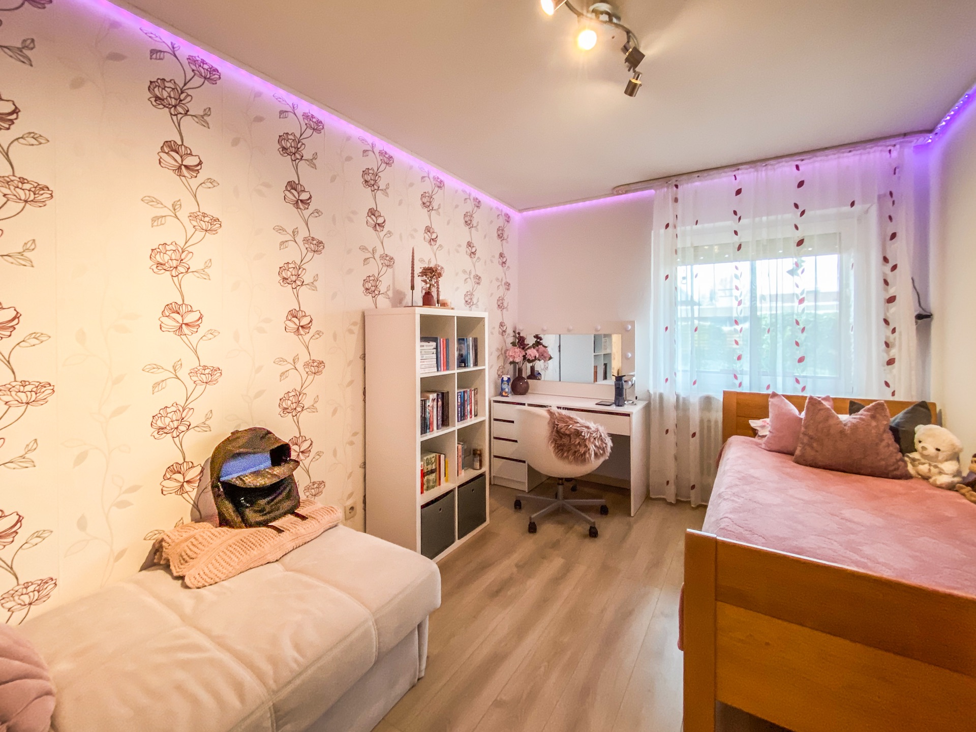 Helles Schlafzimmer mit Blumenmuster-Tapete, Bett, Regal, Schreibtisch und Fenster mit Vorhängen.