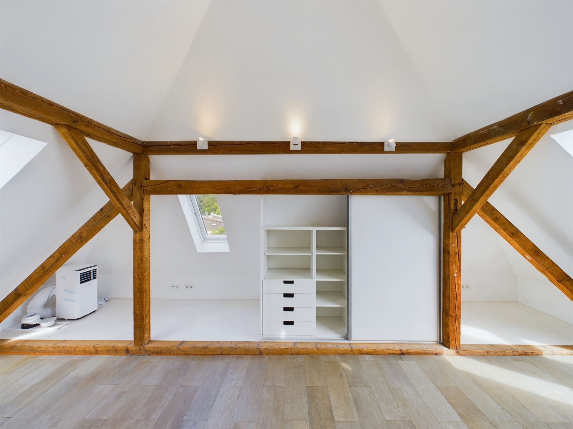 Heller Dachraum mit Holzbalken, Parkettboden und Einbauschrank mit weißen Regalen.