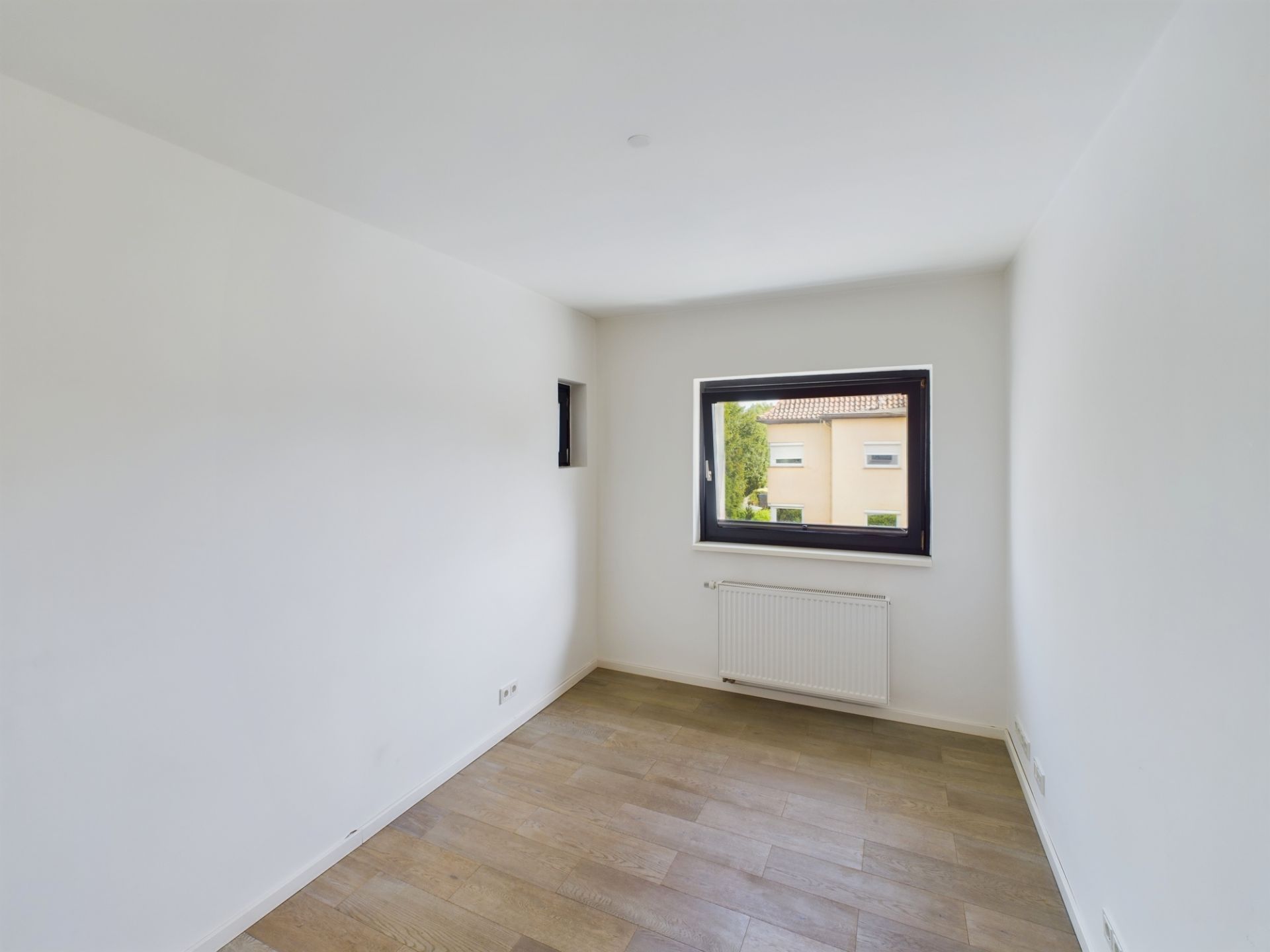 Leerer, heller Raum mit Holzfußboden und großem Fenster, Blick auf Nachbarhaus.