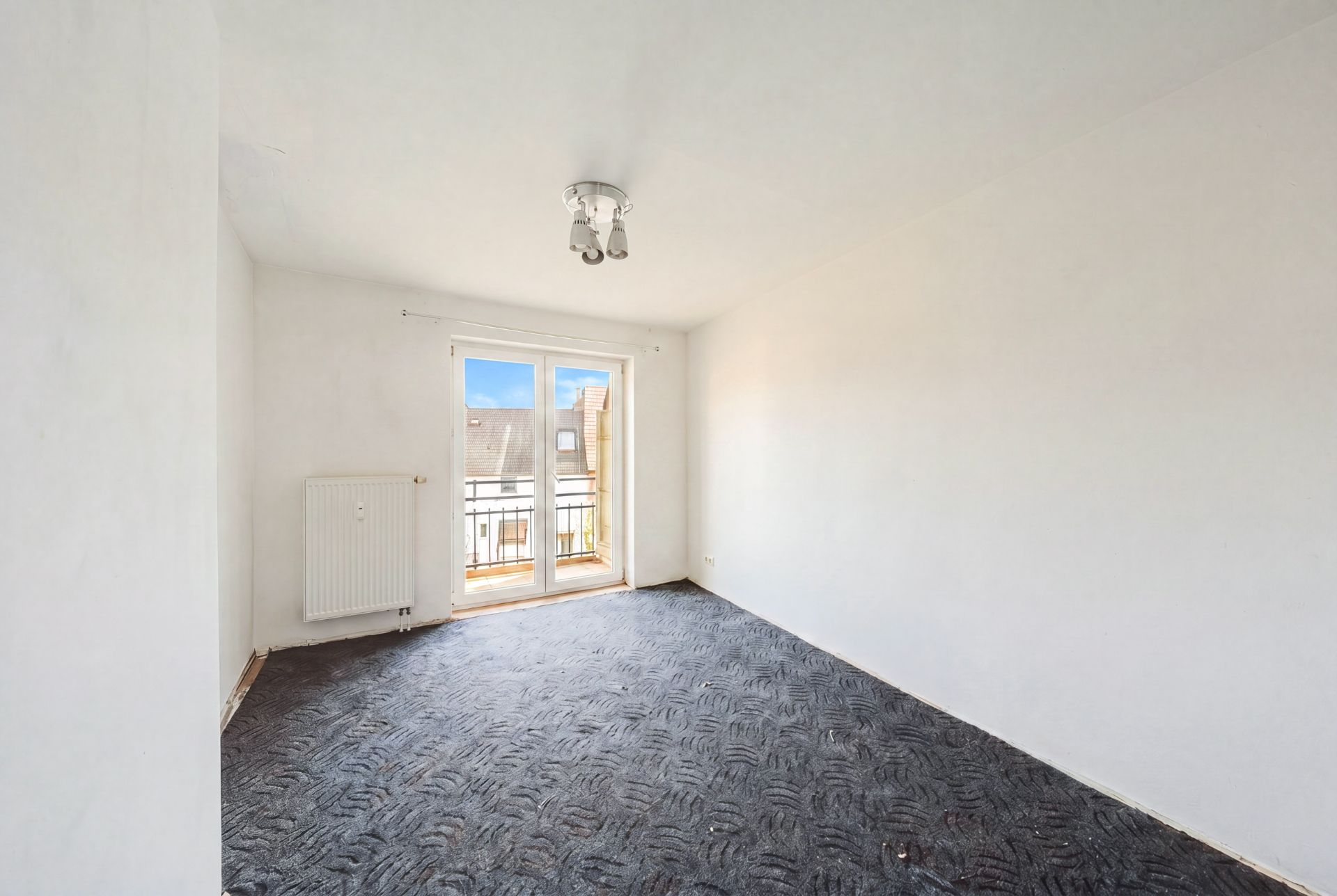 Leeres Zimmer mit Teppichboden, weißen Wänden und Fenstertür zu einem kleinen Balkon.