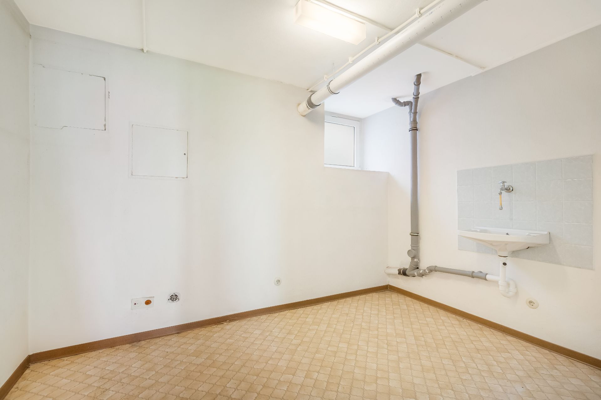 Heller Raum mit Fliesenboden, Waschbecken, Rohren und kleinem Fenster.