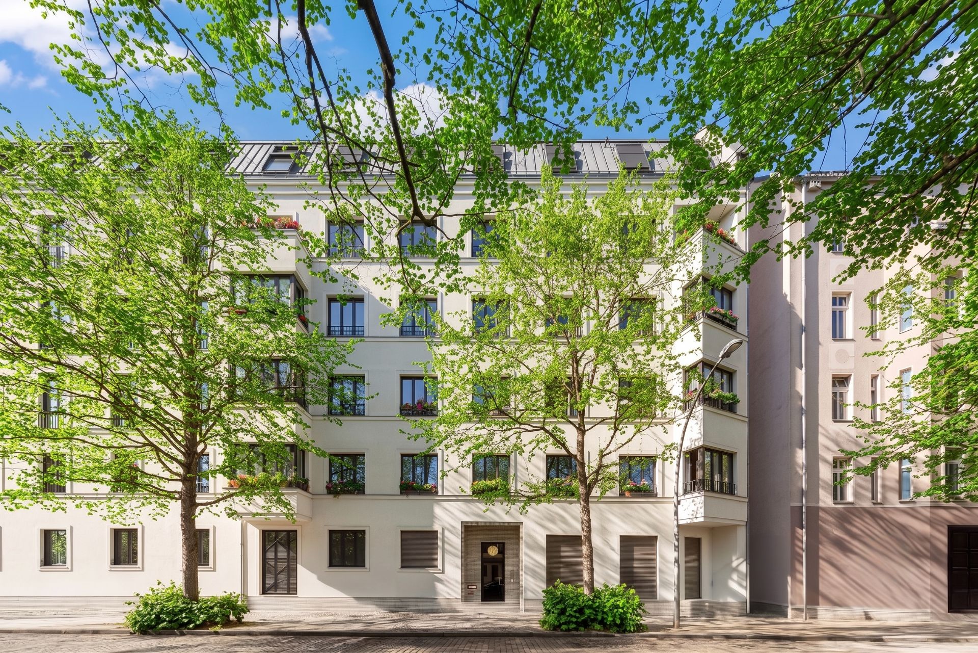 Wohnhaus mit mehreren Fenstern, Bäumen davor und blauem Himmel im Hintergrund.
