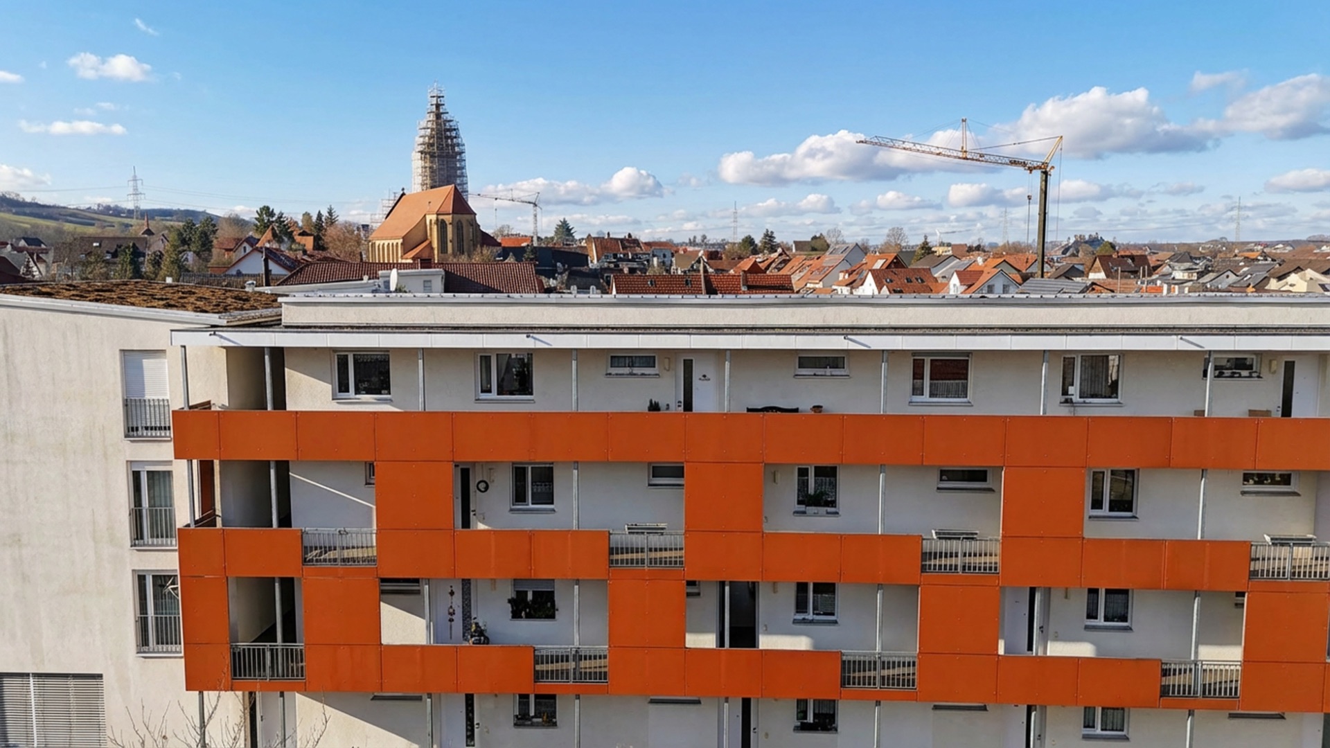 Wohngebäude mit orangen Fassadenelementen und Balkonen, im Hintergrund viele Dächer und eine Kirche.