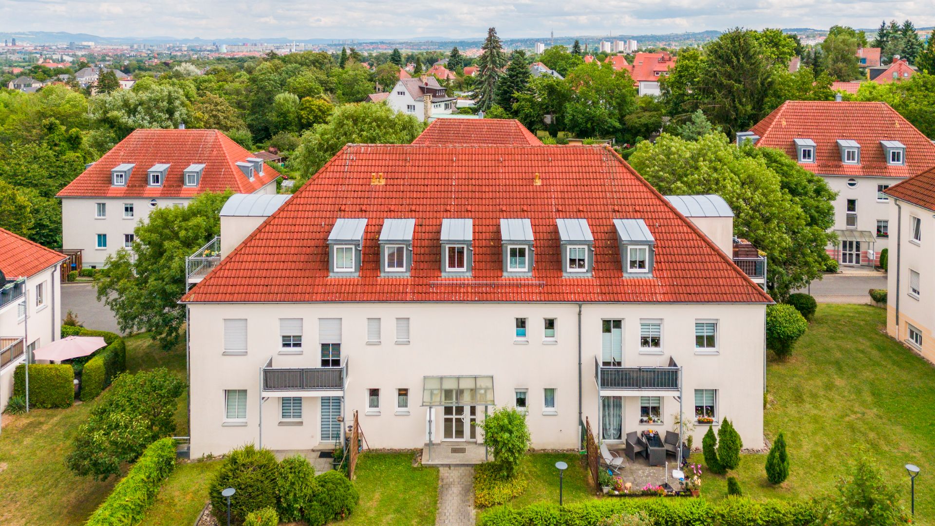 Mehrfamilienhaus mit rotem Dach, weißen Fassaden und mehreren Fenstern, umgeben von Grünflächen.