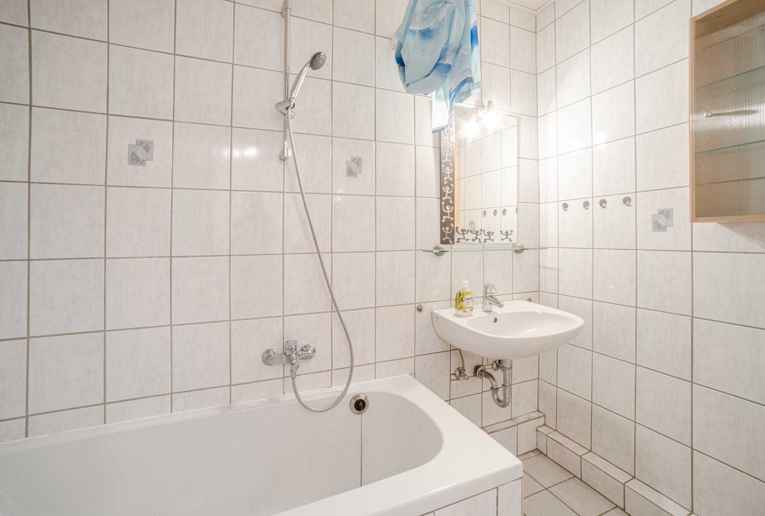 Helles Badezimmer mit Badewanne, Handbrause, Waschbecken und Spiegel, weiße Fliesen an Wand und Boden.