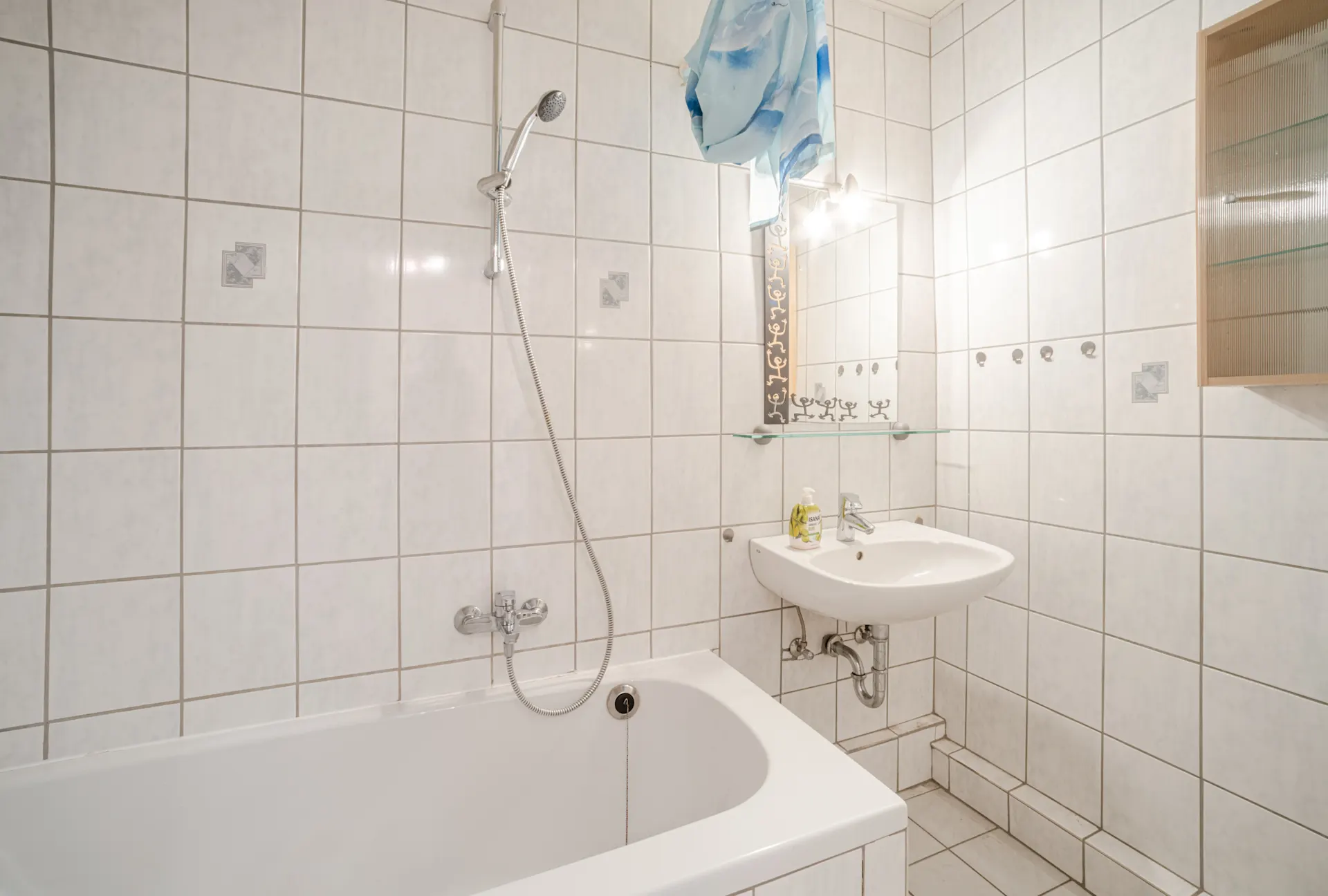 Helles Badezimmer mit Badewanne, Handbrause, Waschbecken und Spiegel, weiße Fliesen an Wand und Boden.
