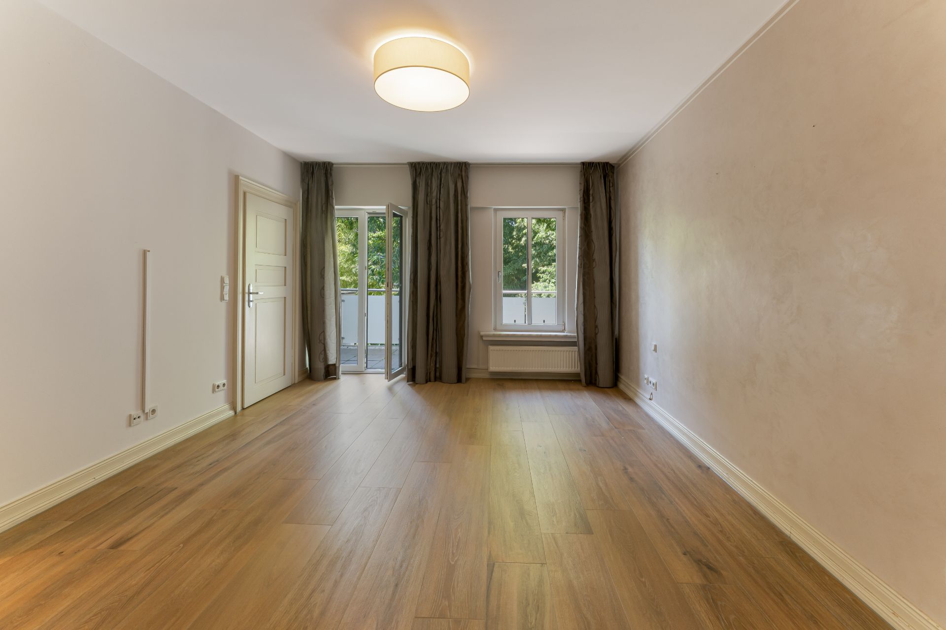 Leerer Raum mit Holzfußboden, großen Fenstern und Vorhängen, Deckenleuchte, helle Wände.