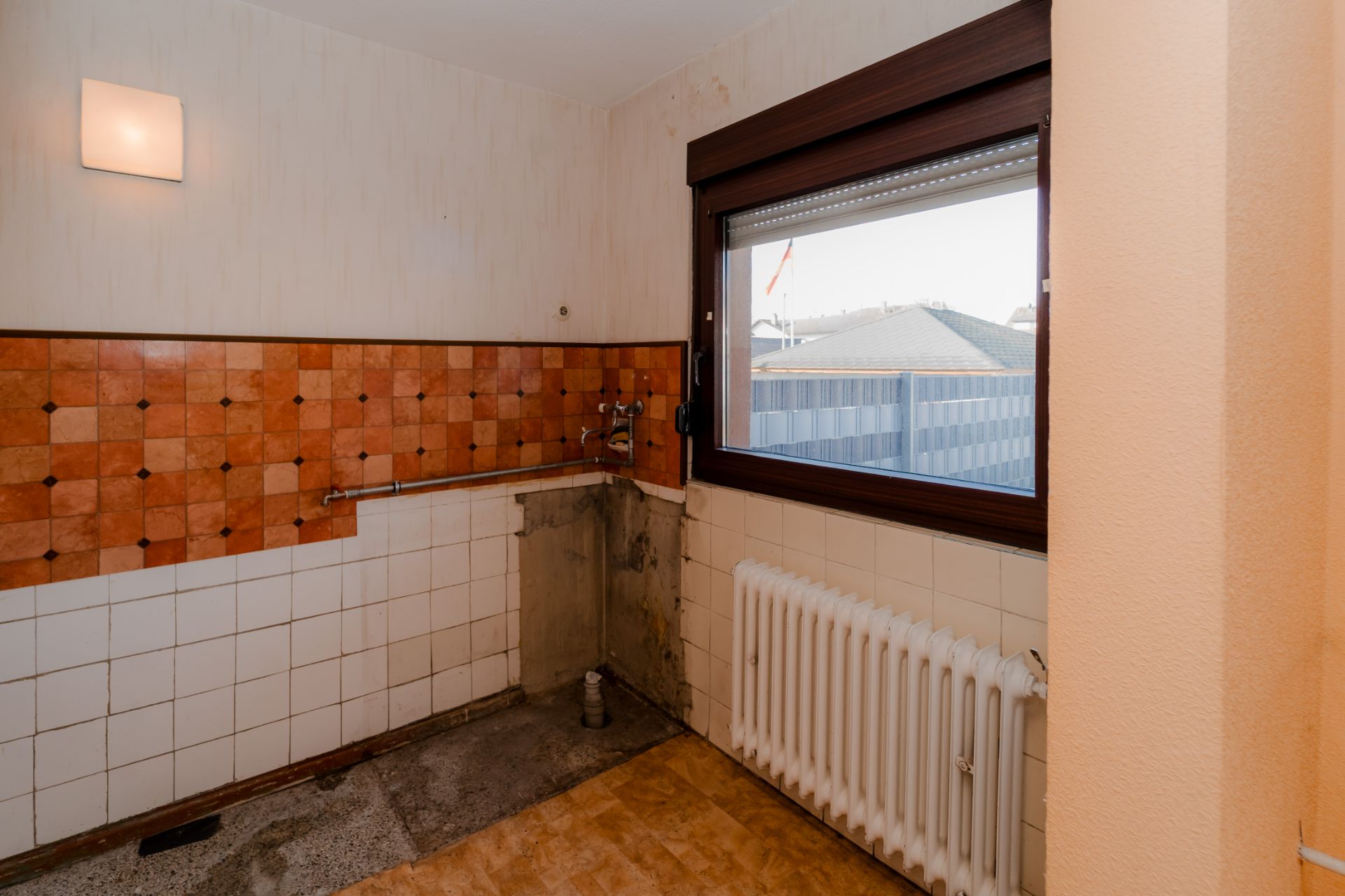Leerer Küchenbereich mit orange-weißen Fliesen, Fenster und Heizkörper.