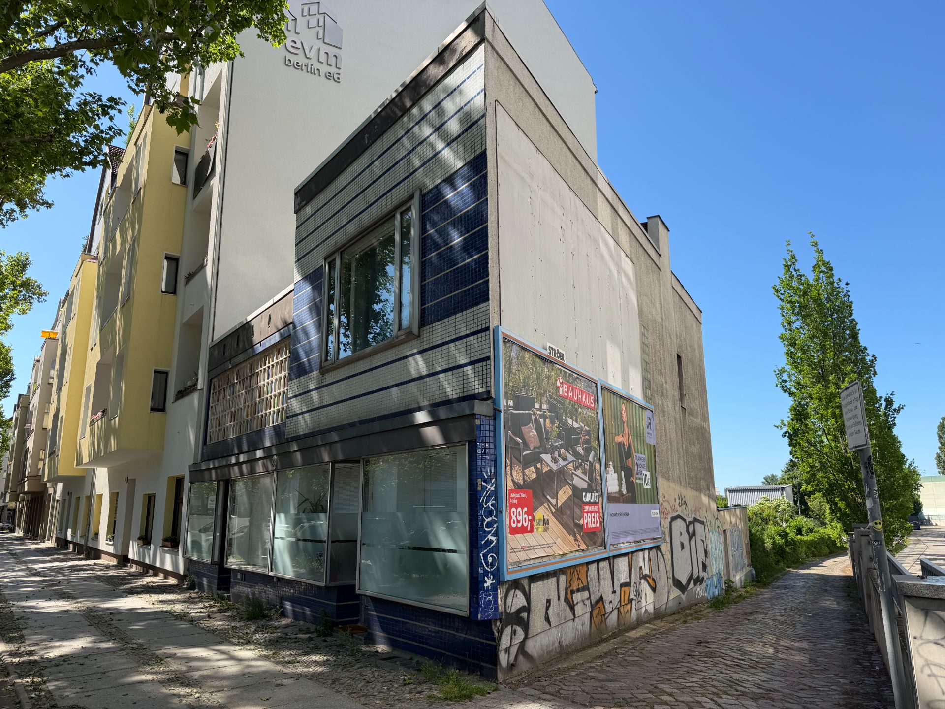 Eckgebäude mit großen Fenstern und Plakatwand, umgeben von Bäumen und Gehweg.