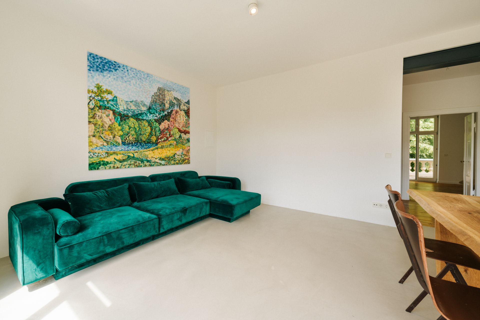 Wohnzimmer mit grünem Sofa, Holztisch, zwei Stühlen und buntem Landschaftsbild an der Wand.