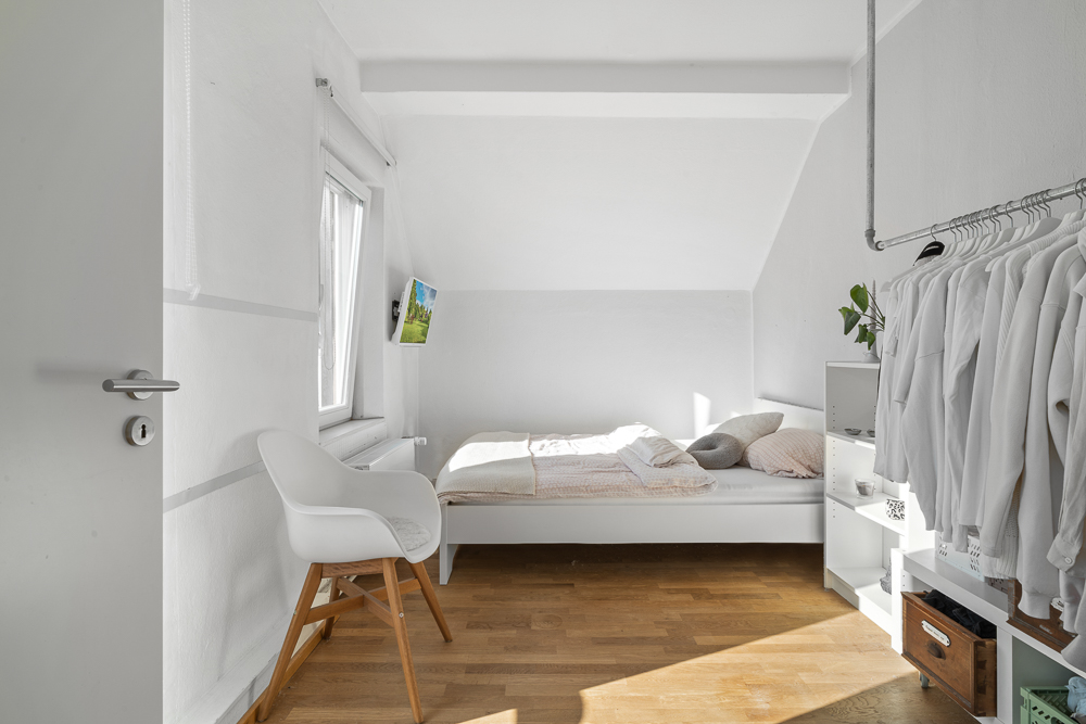 Helles Schlafzimmer mit Einzelbett, Stuhl, Kleiderstange und Holzfußboden.