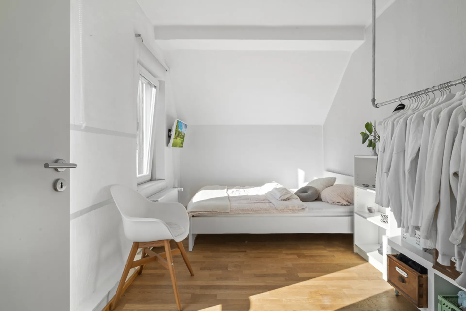 Helles Schlafzimmer mit Einzelbett, Stuhl, Kleiderstange und Holzfußboden.