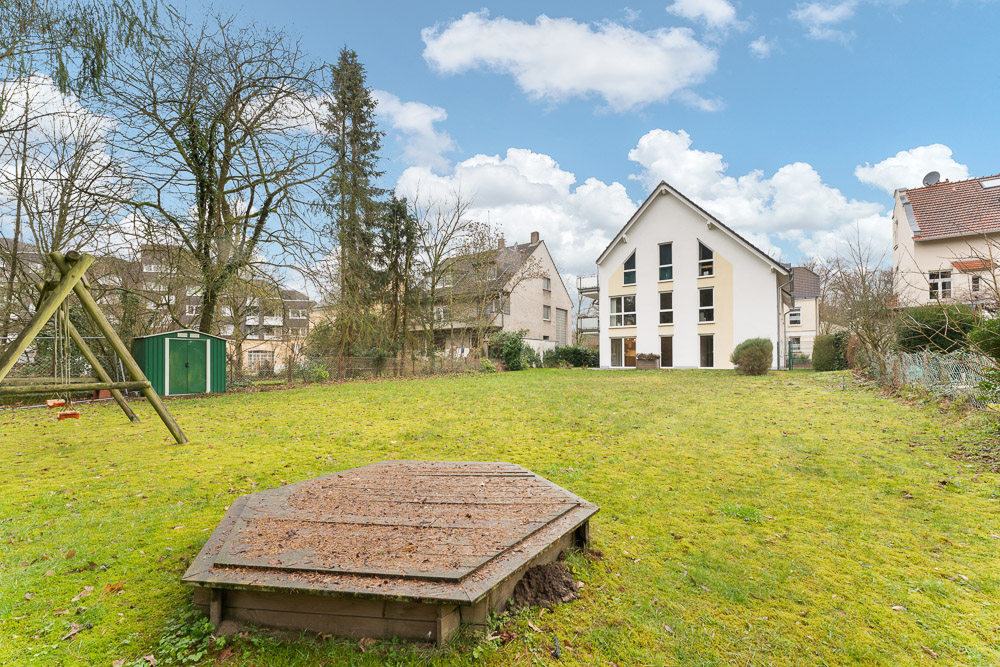 Grüner Garten mit Schaukel, Sandkasten und großem weißem Wohnhaus im Hintergrund.