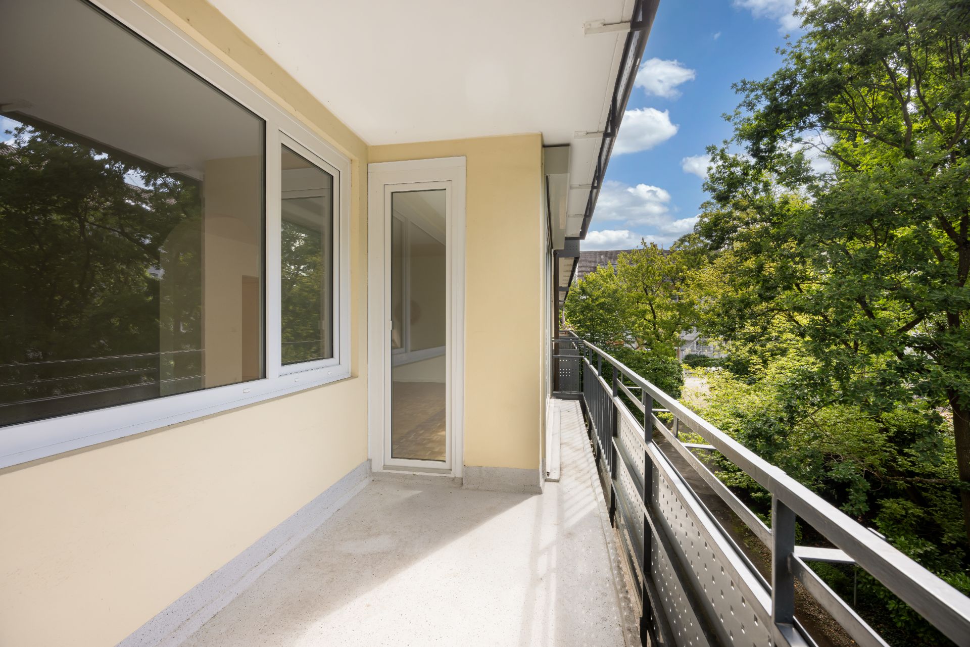 Heller Balkon mit Geländer, großen Fenstern und Blick auf Bäume.