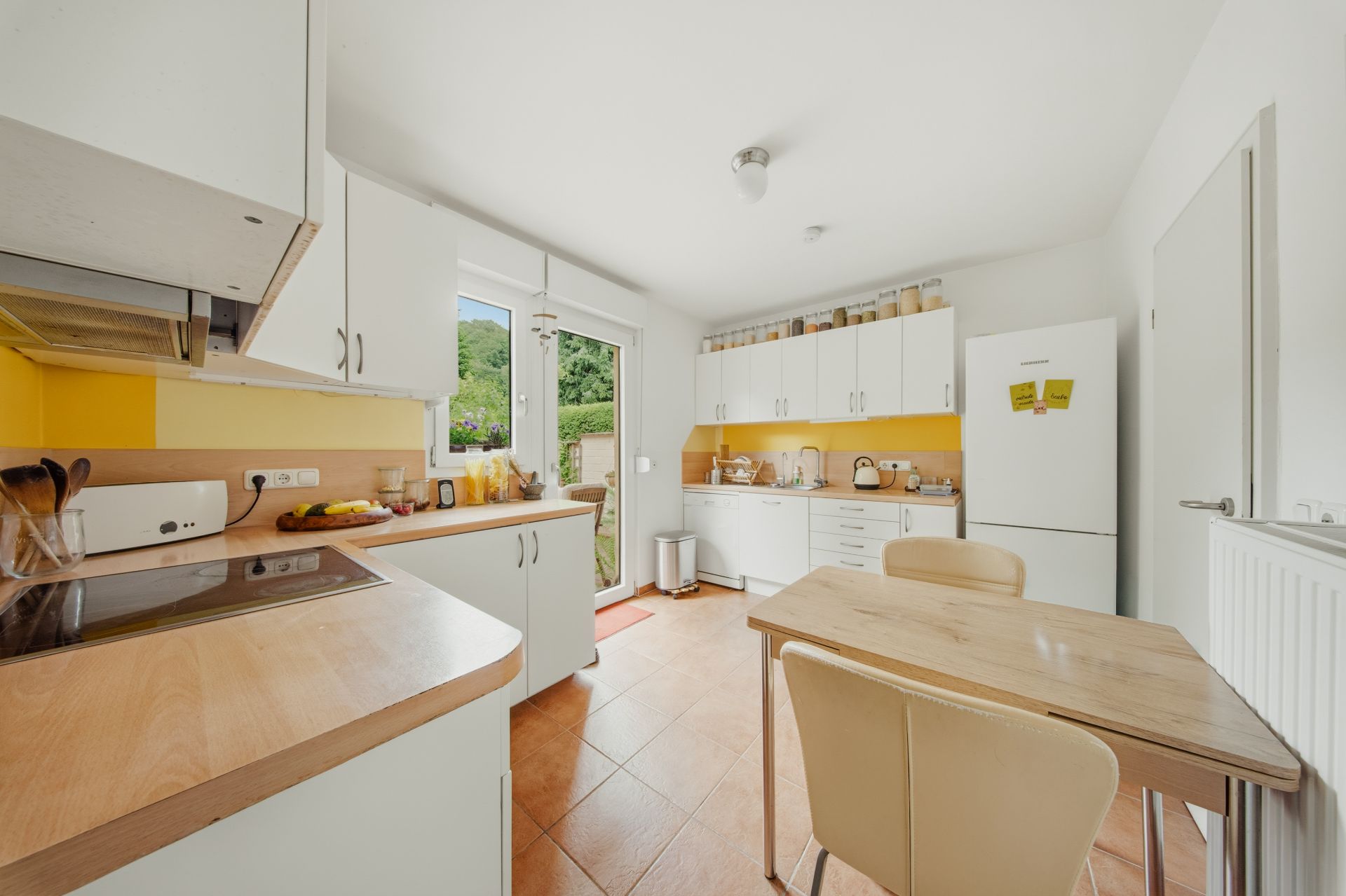 Helle Küche mit weißen Schränken, gelbem Fliesenspiegel, Holztisch und Blick in den Garten.