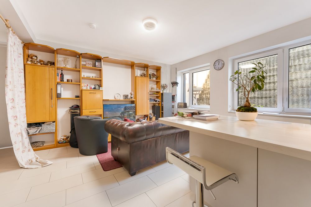 Helles Wohnzimmer mit braunem Sofa, Sessel, Regalwand, Tresen und großen Fenstern.