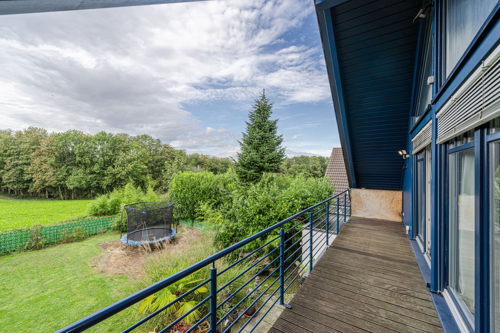 Balkon mit Holzbohlen und blauem Geländer, Blick auf Garten mit Trampolin und Bäumen.