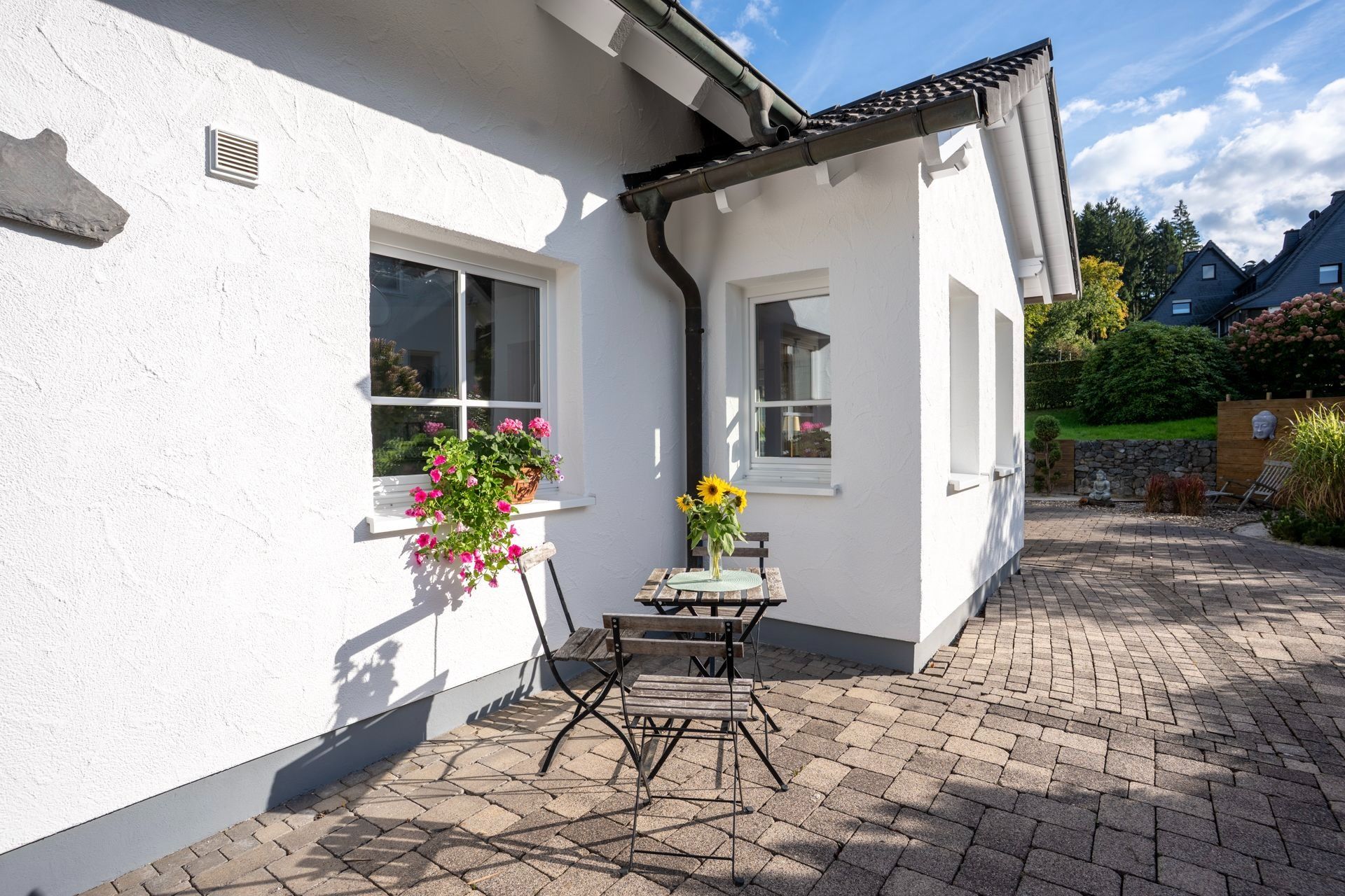 Kleine Terrasse mit Tisch, zwei Stühlen und Blumen vor weißem Haus.